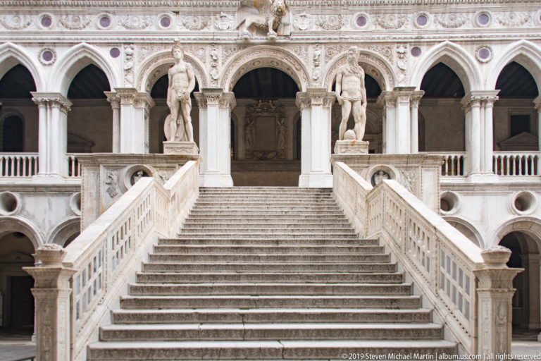 Frontal view up the Scala dei Giganti, with broad marble steps, balustrades, and two colossal statues at the landing.