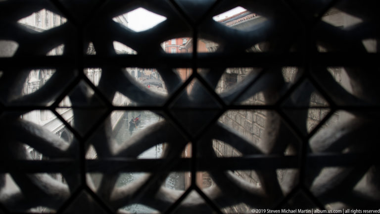 Blurred Venetian exterior seen through a dark geometric grille of diamond and curved openings.
