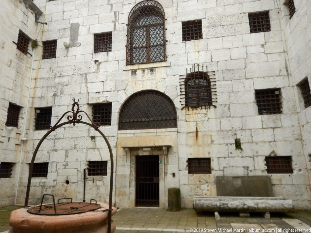 Stone prison courtyard with a circular wellhead, iron frame, barred windows, benches, and a plain doorway.