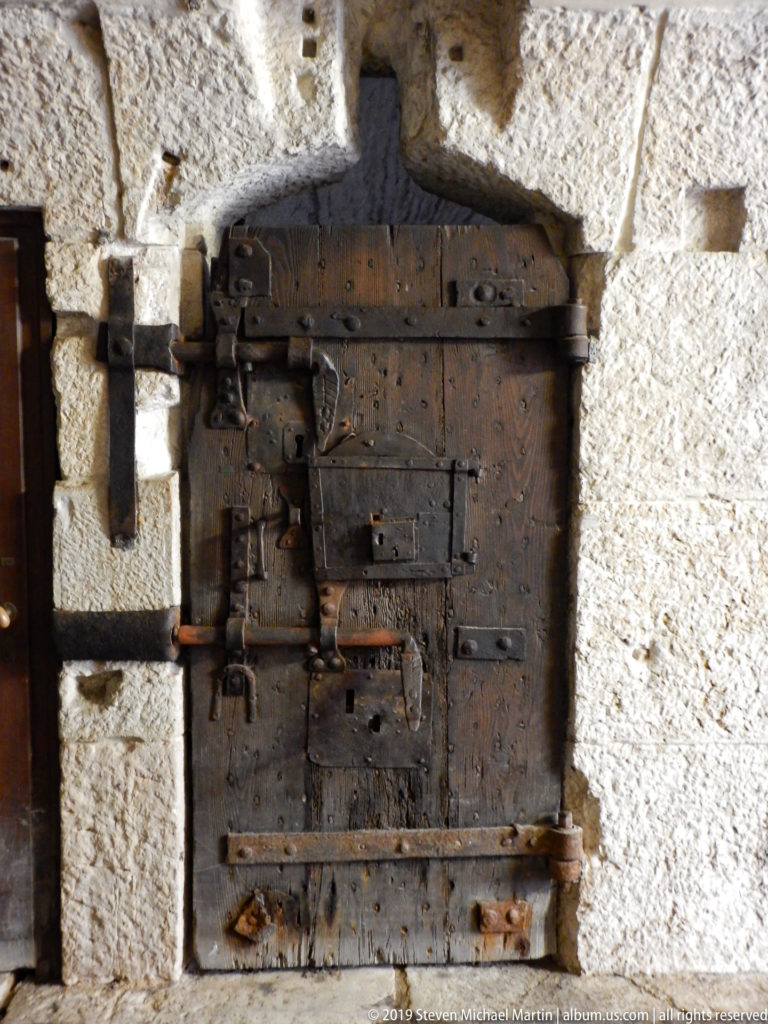 Small heavy wooden door reinforced with iron straps, hinges, studs, and locks in a rough stone wall.