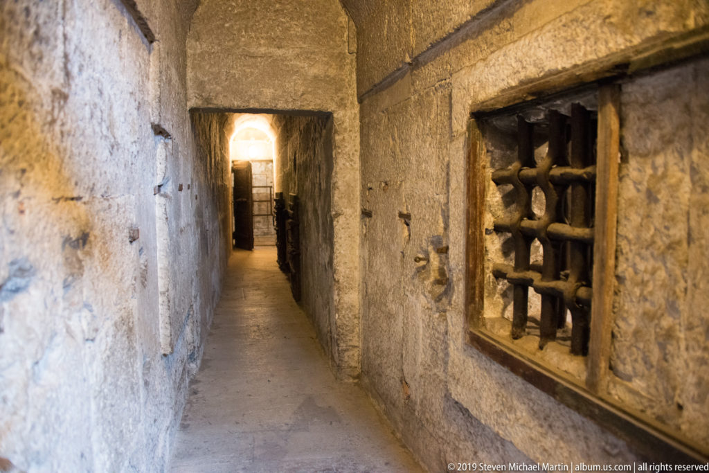 Narrow rough-stone corridor with a heavy barred opening on the right and light visible at the far end.