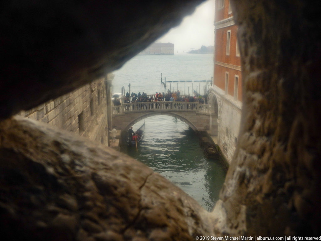 Canal, pedestrian bridge, and waterfront buildings seen through a dark, narrow stone opening from inside the palace complex.