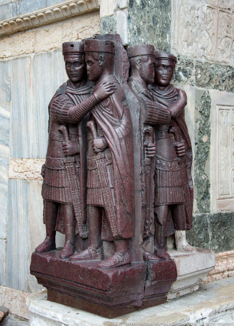 Dark red porphyry sculpture of four armored rulers embracing in pairs around the corners of a block.