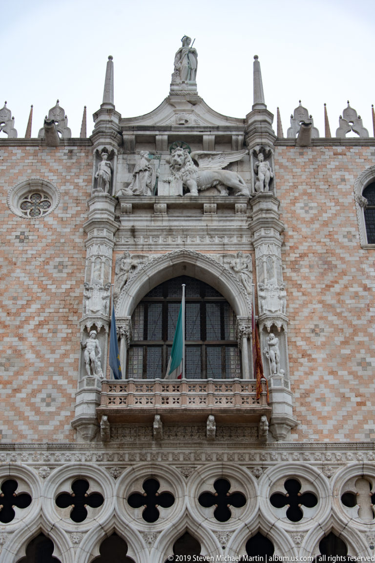 Sculpted stone balcony and arched window set into patterned brickwork above a row of Gothic quatrefoils.