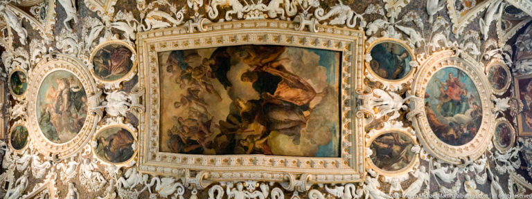Oblique view of a decorated ceiling with a central rectangular painting, oval scenes, pale relief ornament, and gilded borders.