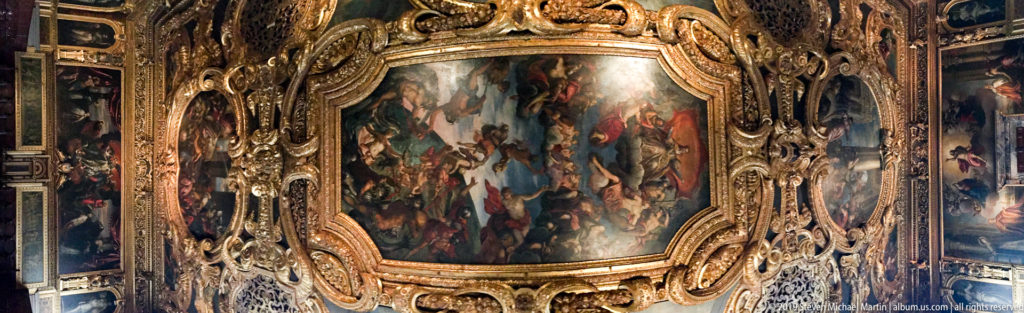 Large dark narrative painting surrounded by a deep gilded carved frame, with additional paintings continuing along the wall.