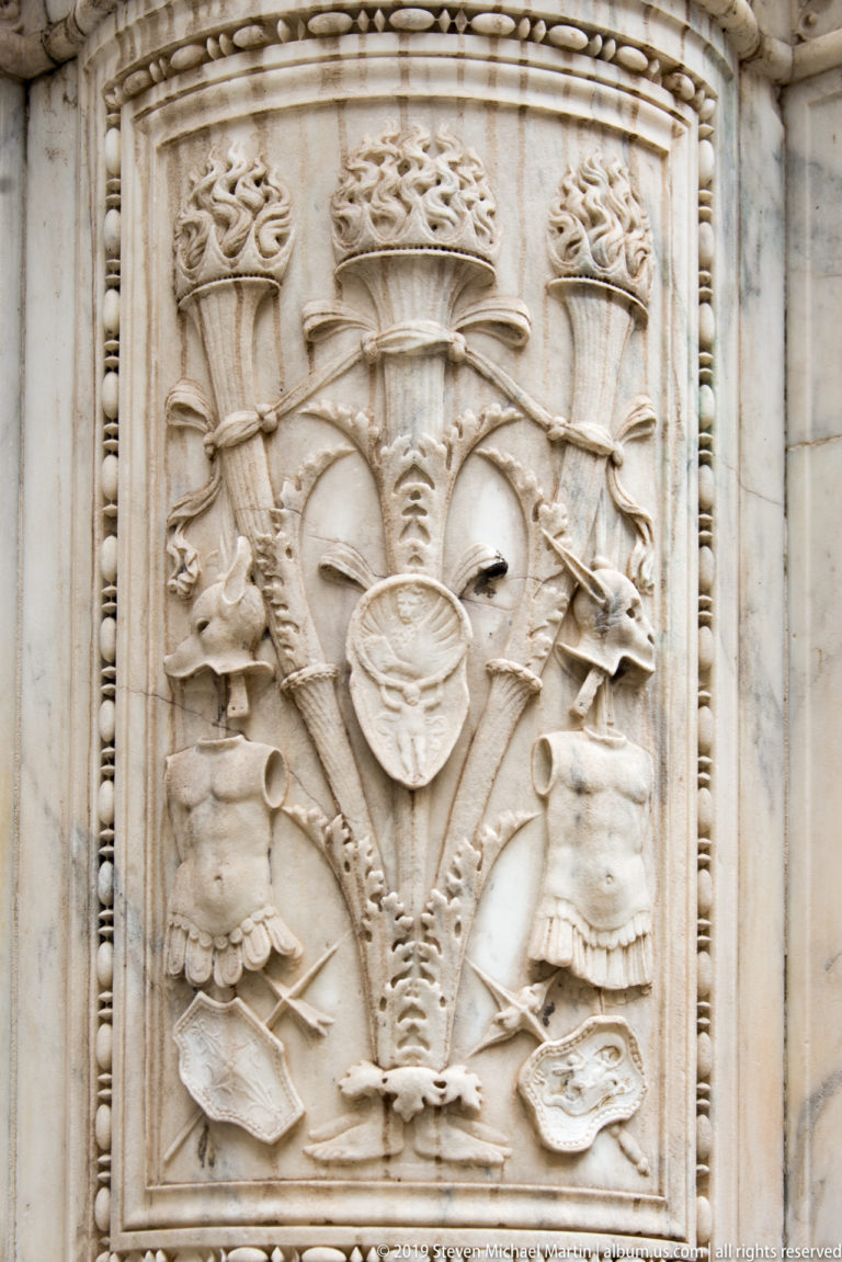 Rectangular stone relief of helmets, shields, standards, armor, and other military trophies arranged symmetrically.
