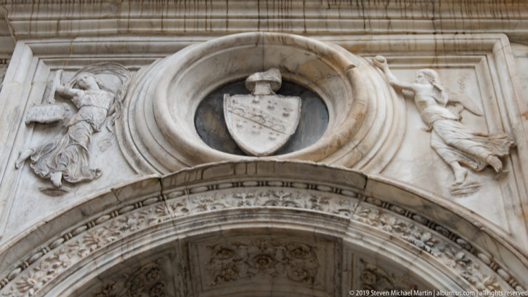 Circular marble frame with a shield above an ornate arch, flanked by carved relief figures.