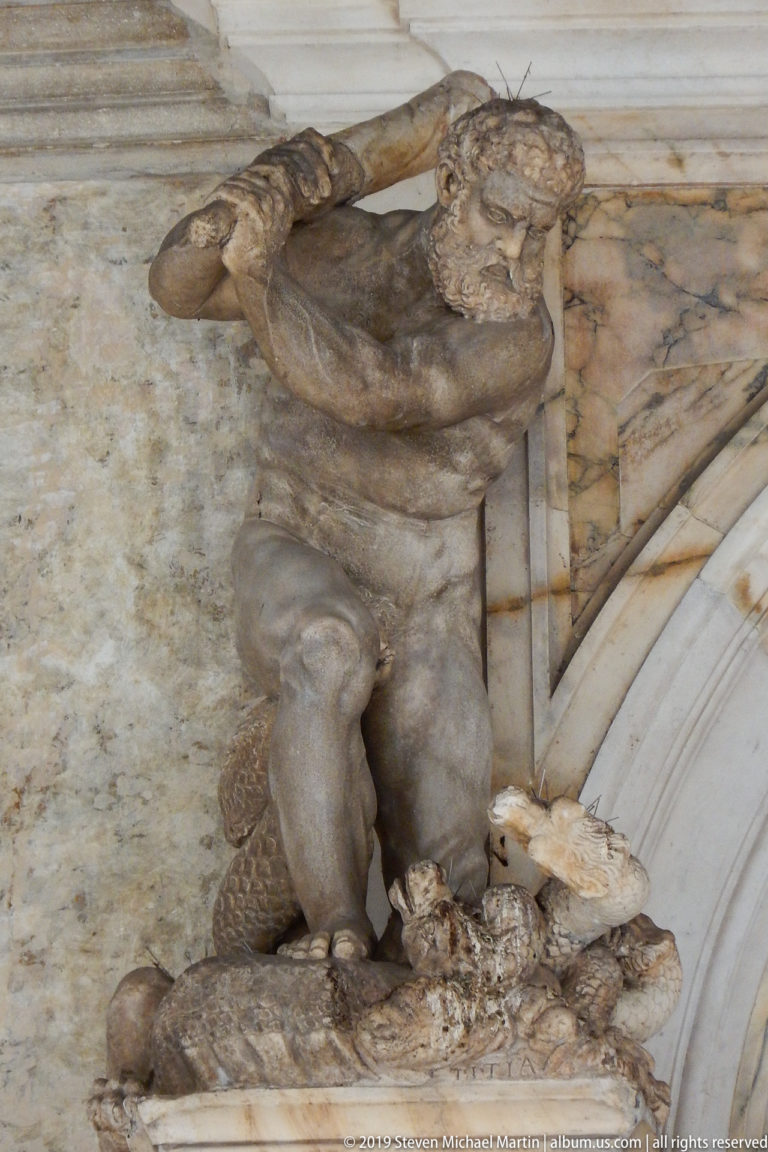 Weathered crouching male figure above an arch, with one arm raised behind his head and rocky forms below.