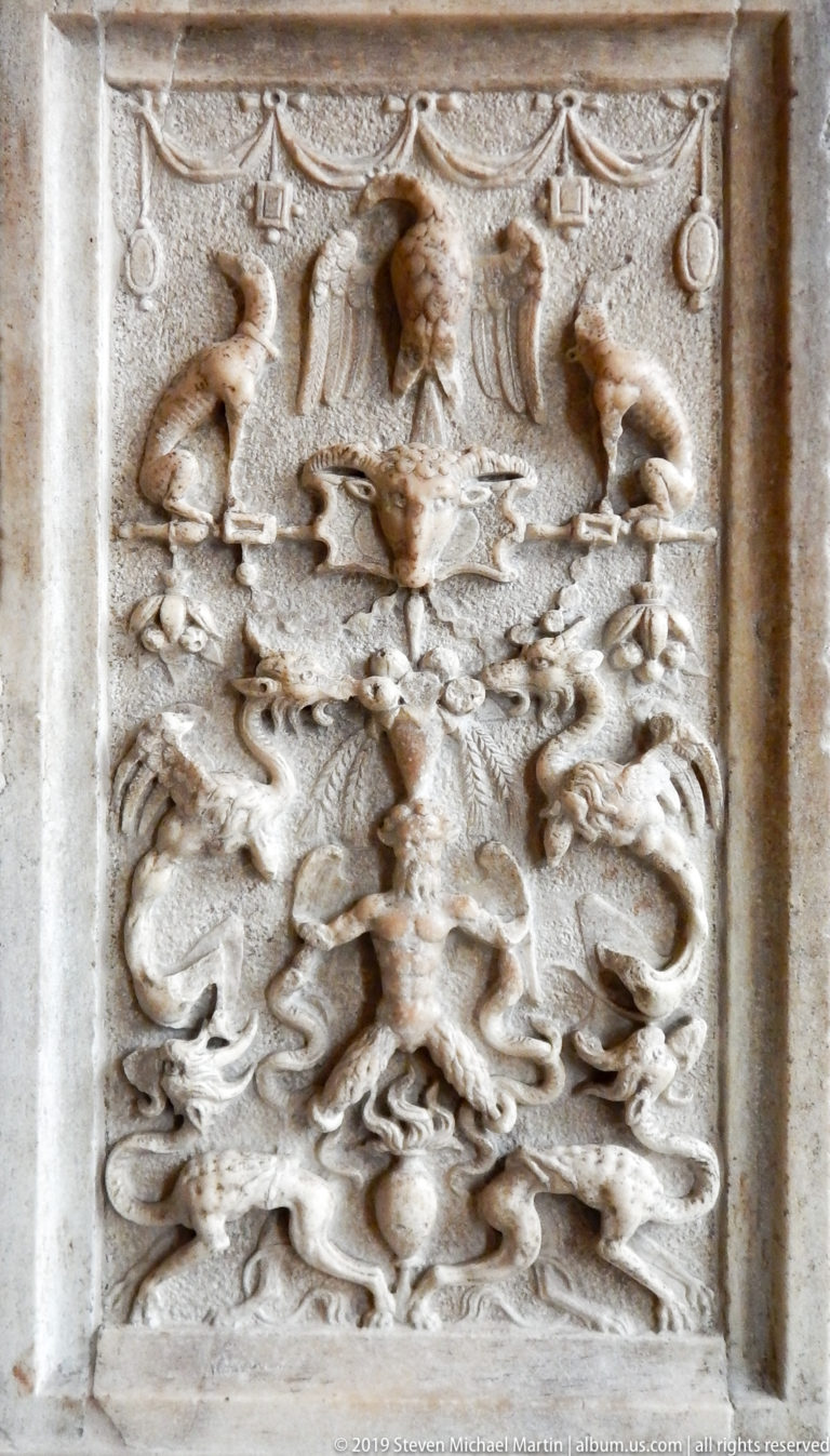 Weathered rectangular stone relief crowded with human, animal, and foliate forms around a central grotesque design.