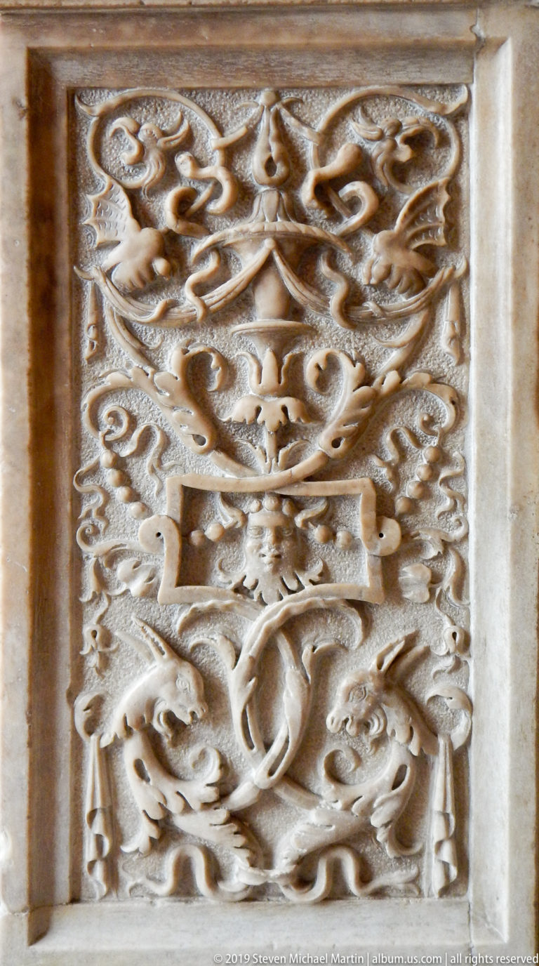 Rectangular pale-stone relief with a central grotesque face, curling foliage, small creatures, and a carved border.