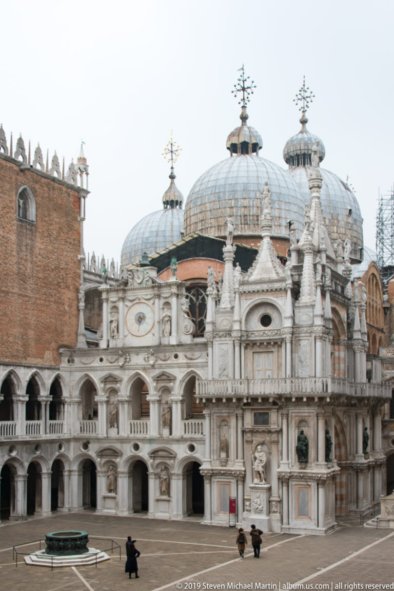 Arco Foscari centered in the palace courtyard with St Mark’s domes, arcades, paving, and a wellhead around it.