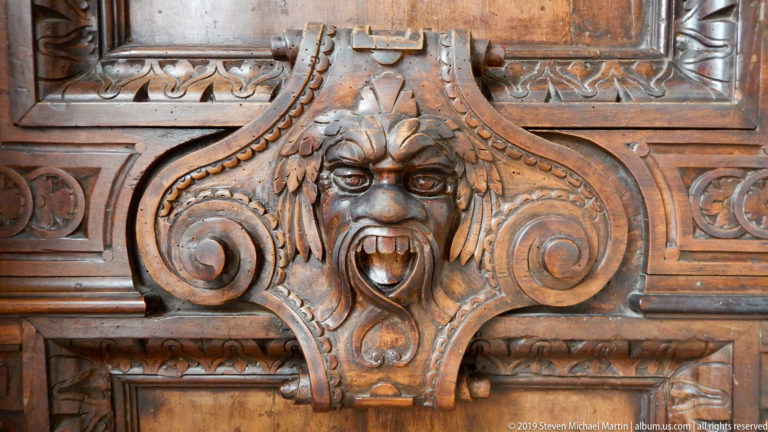 Horned carved wooden grotesque with open mouth and teeth, centered between large spiral volutes.
