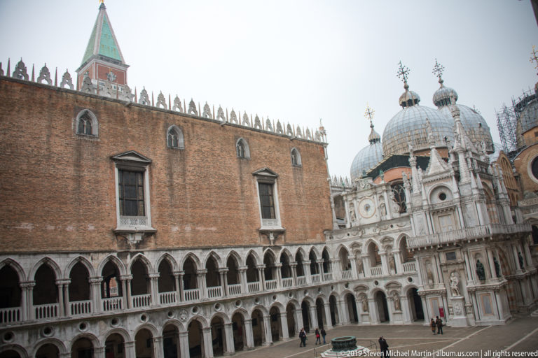 Wide Palazzo Ducale courtyard with brick wing, stacked arcades, Arco Foscari, domes, visitors, and a wellhead.