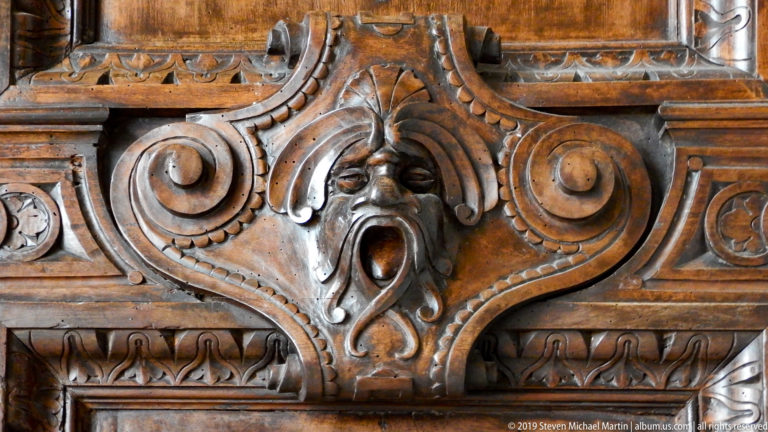 Frontal carved wooden grotesque with open mouth, pointed crest, symmetrical scrolls, and ornamental borders.