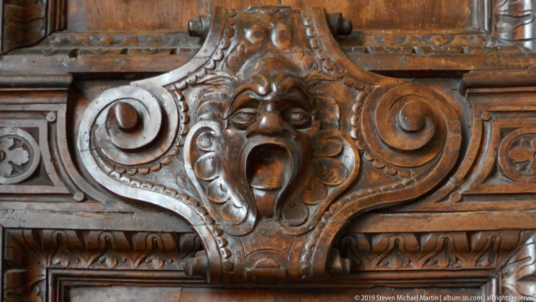 Dark carved wooden grotesque mask with open mouth, framed by large scrolls and leaf-pattern borders.