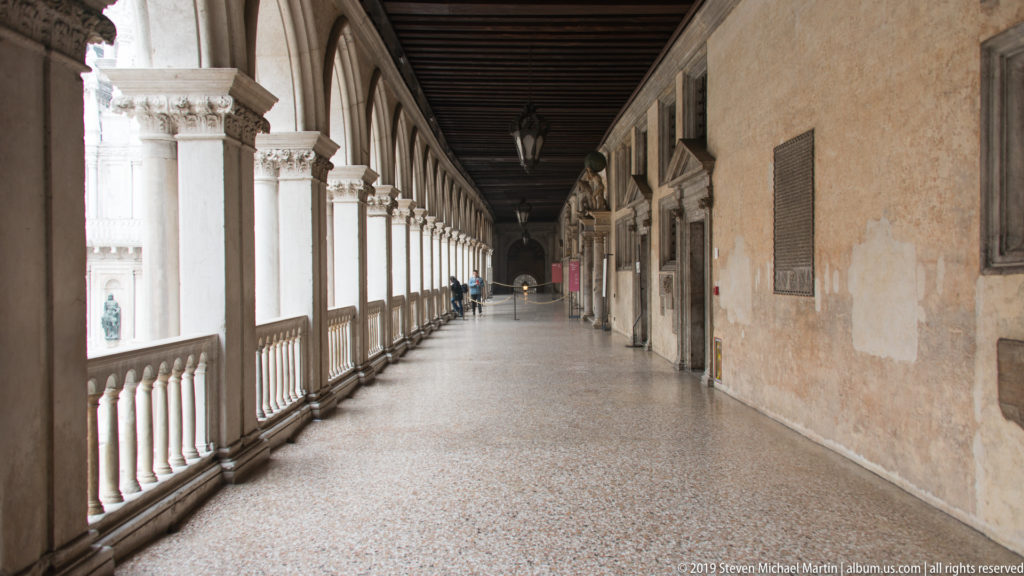Long palace loggia with repeating columns and arches, terrazzo floor, dark ceiling, and a few distant visitors.