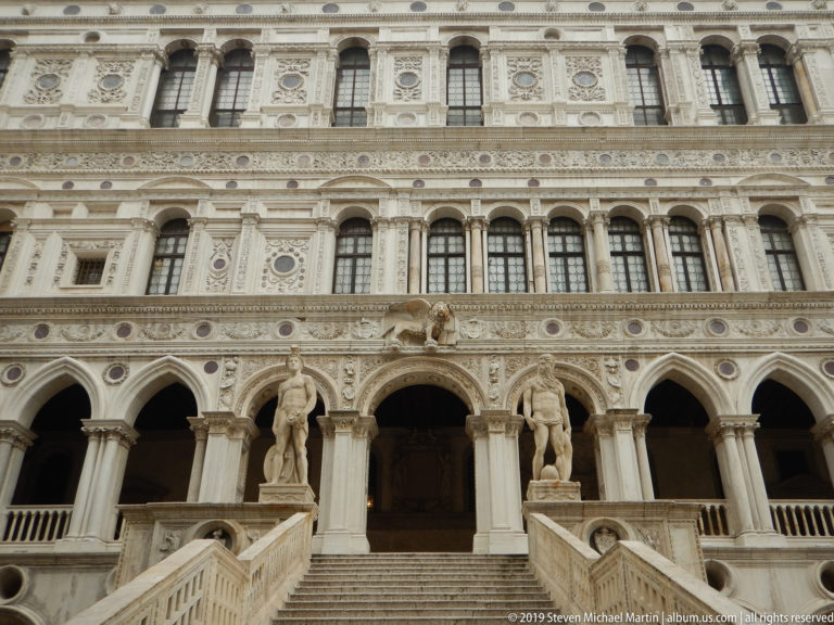 Courtyard façade with broad staircase, paired statues, arcaded loggia, and richly carved upper stories.