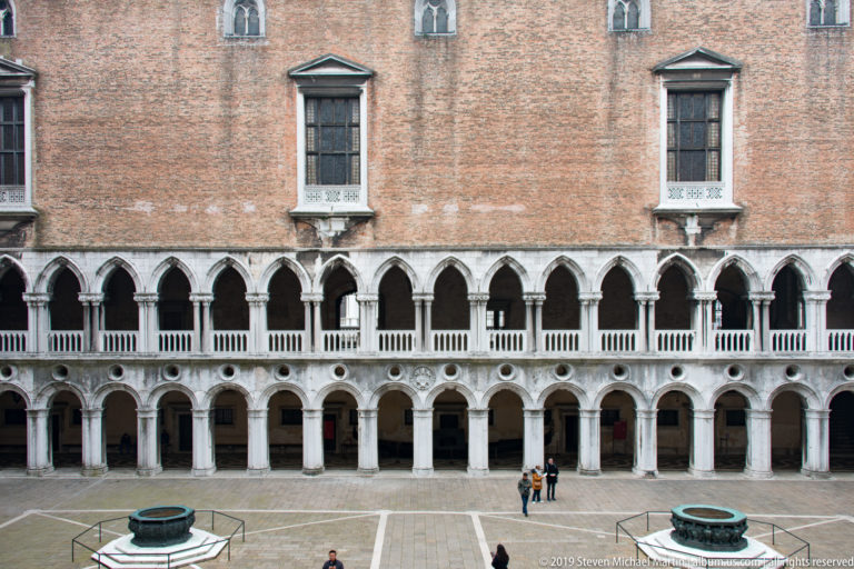 Wide courtyard view with two levels of pale stone arcades, brick walls, two wellheads, and a few visitors.