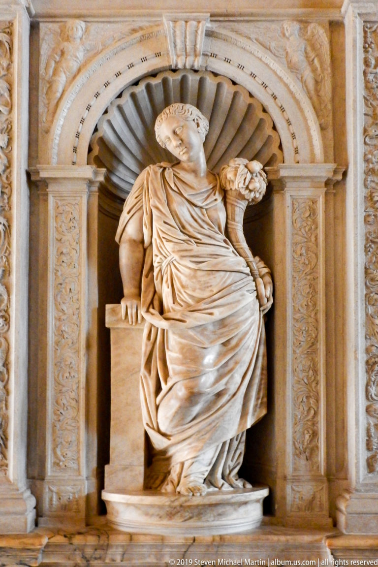 Seated draped marble figure in a scallop-shell niche framed by carved pilasters and ornamental borders.