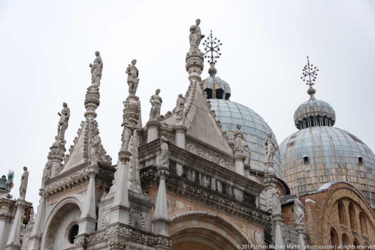 Upper pinnacles and statues of the Arco Foscari set against the domes of St Mark’s Basilica.