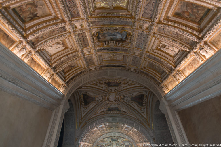 Long vaulted passage with painted coffers, sculptural ceiling ornament, and an illuminated arch at the far end.