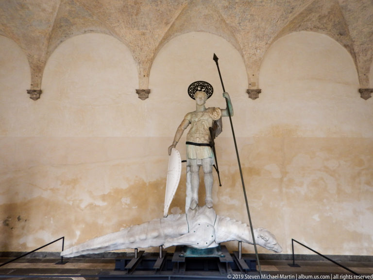 Armored saint holding a long spear and shield stands on a large pale reptilian creature beneath vaulted arches.