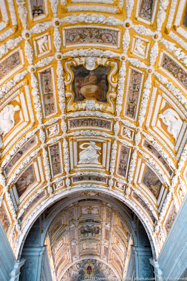 Upward view of a coffered barrel vault with alternating painted scenes, pale relief panels, and gold-edged geometric frames.