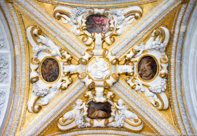 White-and-gold ceiling compartment centered on a pale mask, with oval paintings, scrolls, and small modeled figures around it.