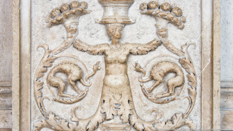 Pale symmetrical relief with a central grotesque figure, paired animal forms, curling foliage, and a small urn above.