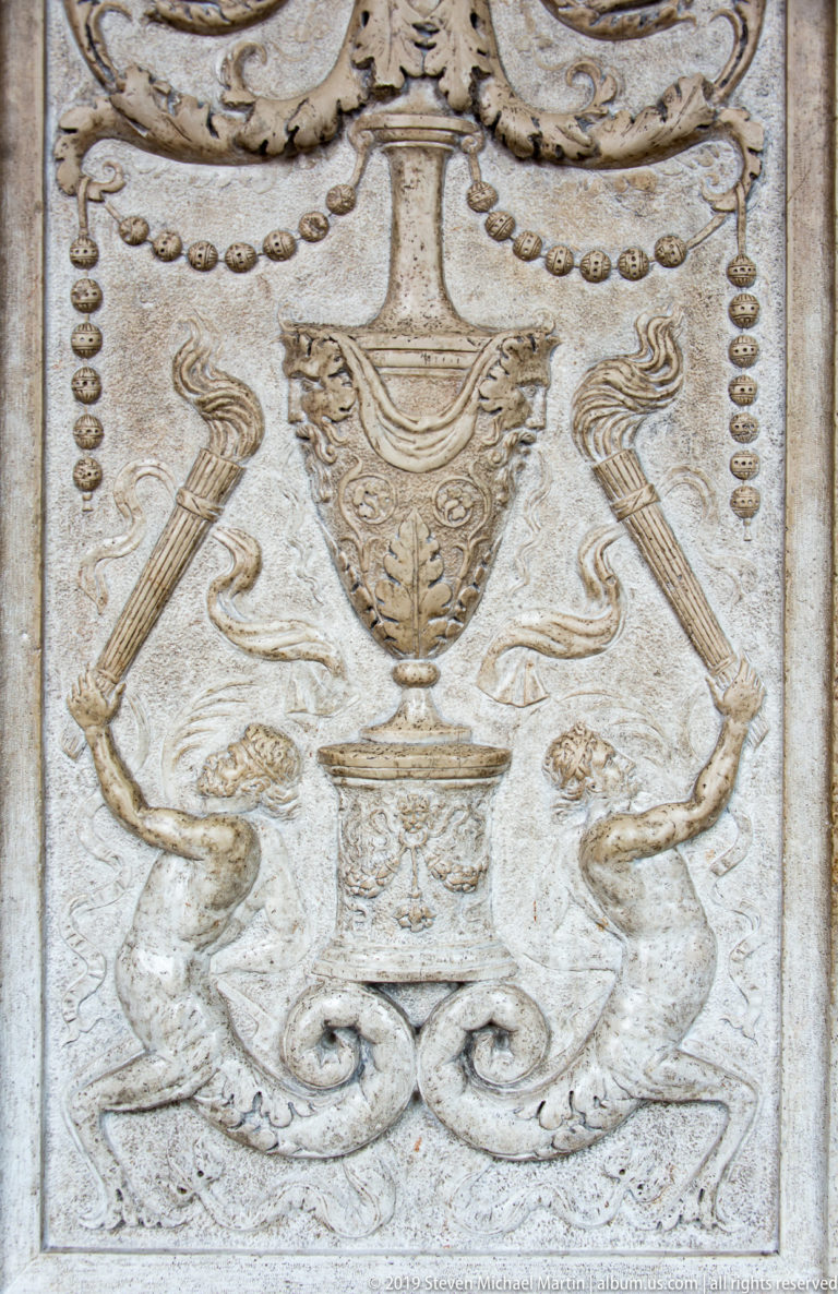 Pale carved stone panel with a central urn, paired hybrid creatures, scrolls, and hanging bead motifs.