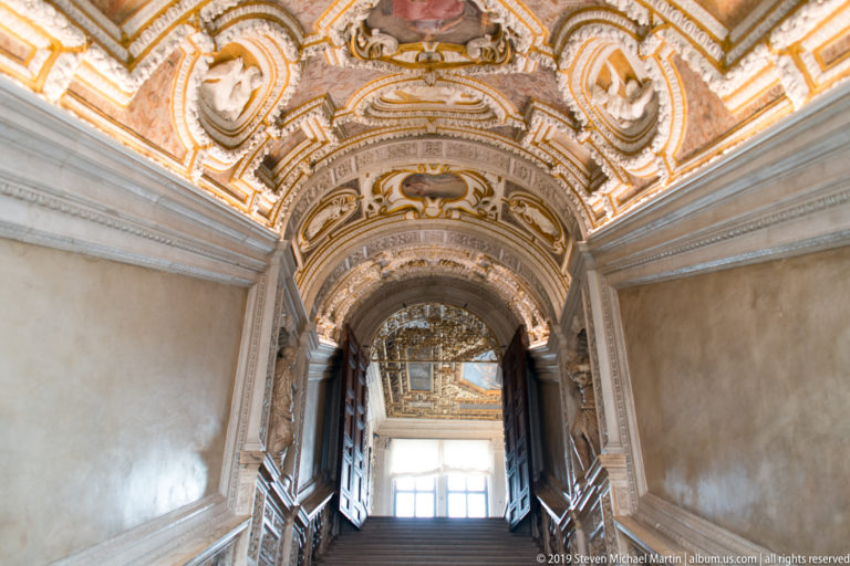 Stair corridor rising beneath a richly painted barrel vault toward a bright windowed landing, with dark open doors along the sides.