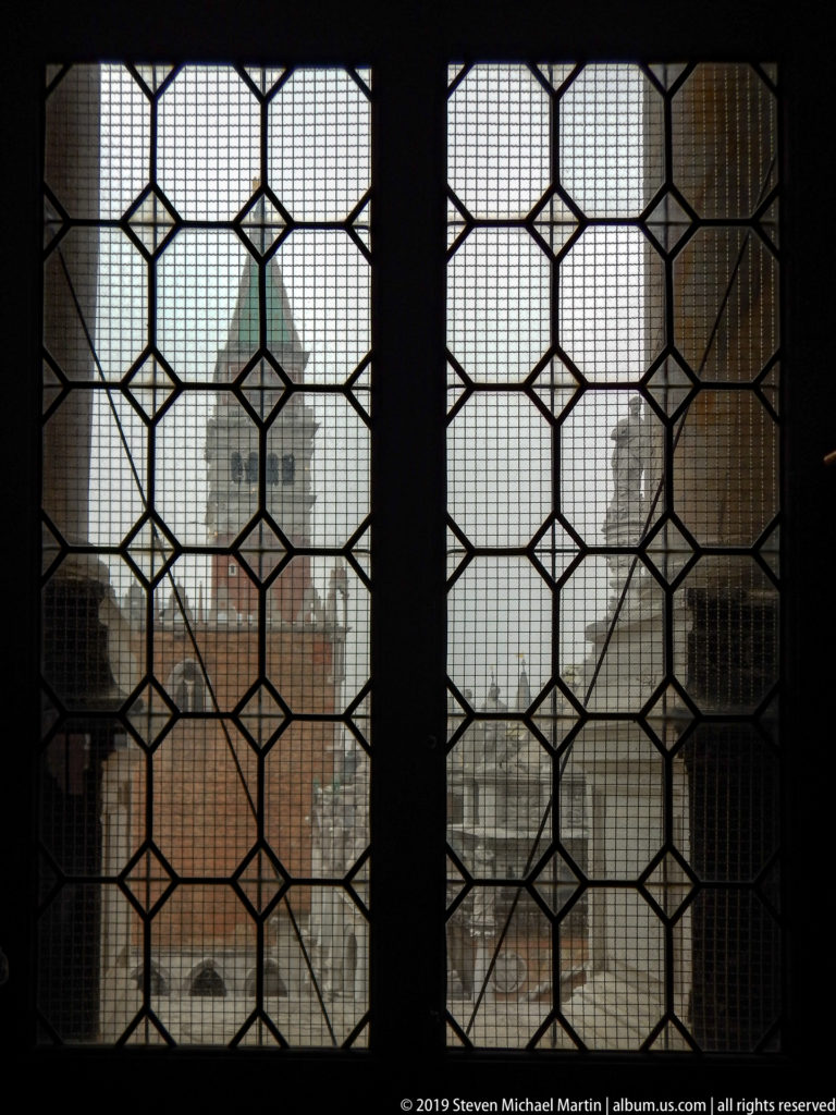 Dark geometric leaded window framing a hazy view of rooftops and towers across Venice.