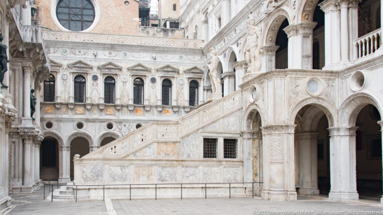 Side view of an ornate marble staircase crossing a courtyard lined with arches, columns, and carved stone galleries.