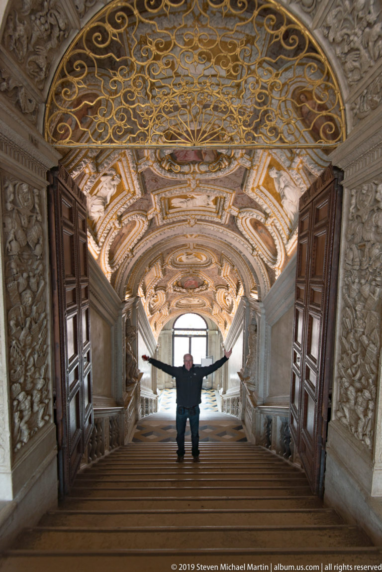 Visitor with arms outstretched on a broad ornate staircase beneath a gilded grille and richly decorated barrel vault.