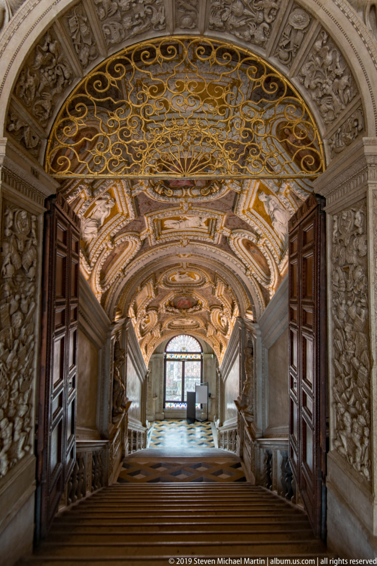 Broad palace staircase beneath an ornate barrel vault, with open wooden doors, relief-covered walls, and a gilded metal grille overhead.