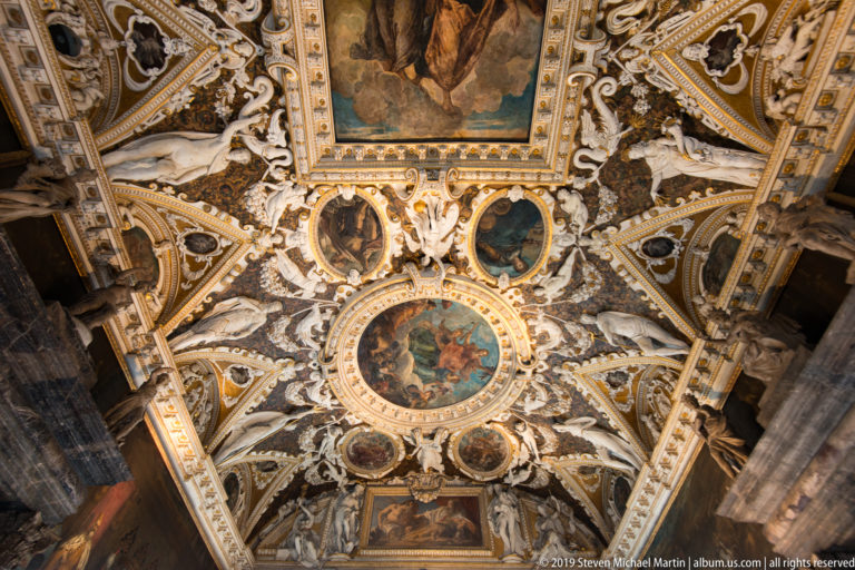 Oblique view along an ornate vault with rectangular and circular paintings, white stucco figures, and gilded dividing bands.
