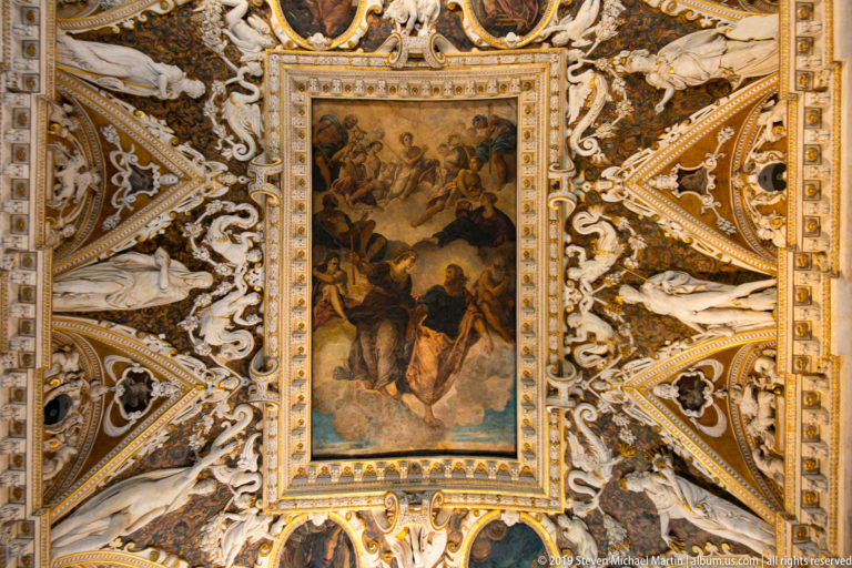 Rectangular multi-figure ceiling painting surrounded by white sculptural stucco, gilded ribs, scrolls, and triangular compartments.