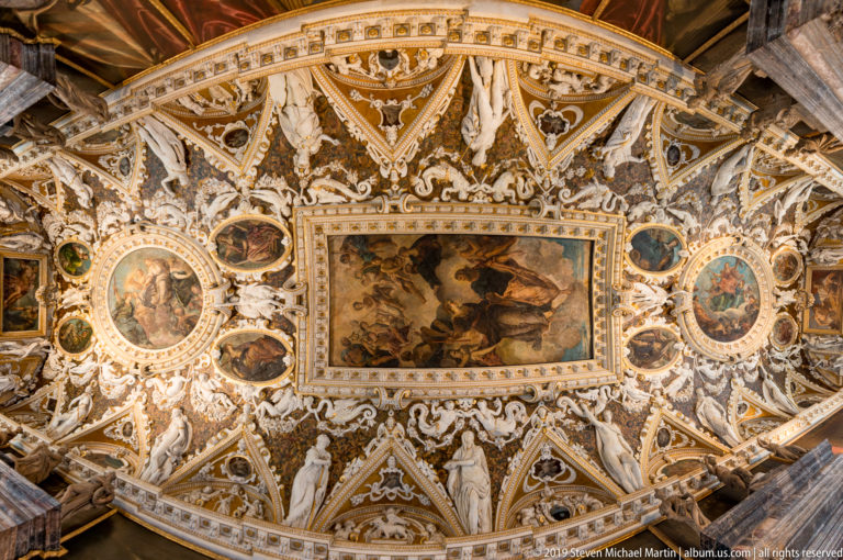 Elaborate white-and-gold vaulted ceiling with a central rectangular painting, oval medallions, sculpted figures, and pointed compartments.