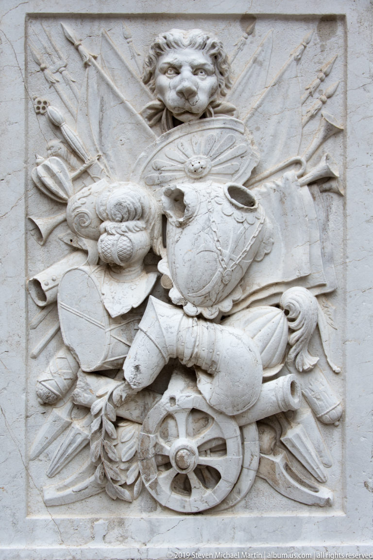 Rectangular stone relief filled with a helmeted head, shields, armor, spears, standards, and a wheel.