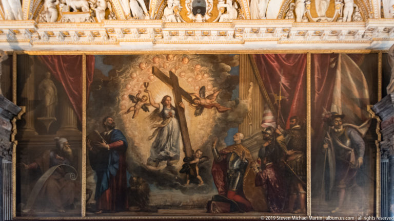 Large sacred painting with a radiant central figure, prominent cross, angels, attendants, red drapery, and dark side figures.