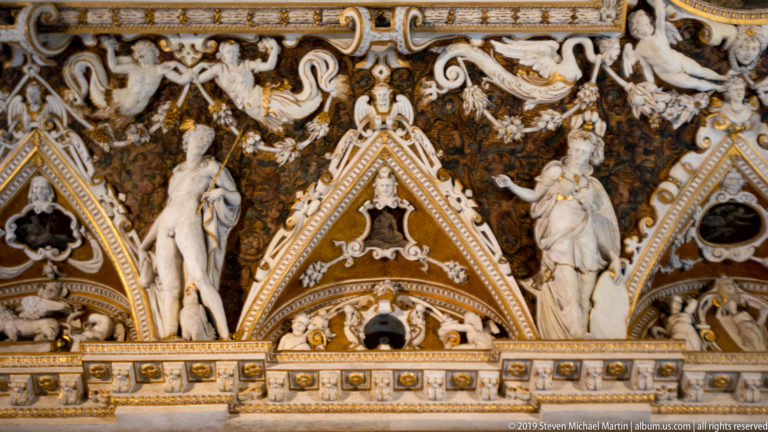 Two pale standing female sculptures flanking a pointed ceiling compartment amid dense white scrollwork and gold architectural trim.