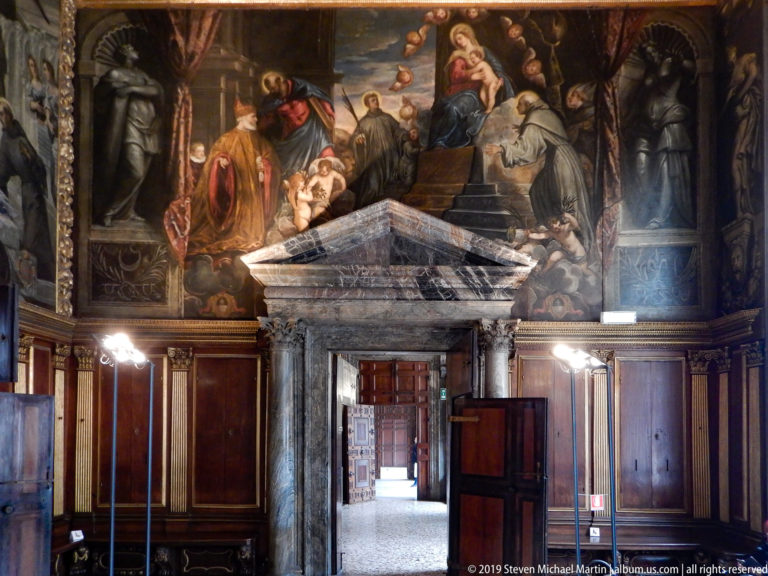 Gray stone doorway in dark wood paneling beneath a large multi-figure painting, with an open view into adjoining rooms.