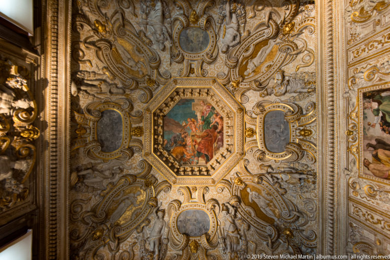 White-and-gold ornamental ceiling with an octagonal central painting, gray oval panels, scrollwork, masks, and modeled stucco.