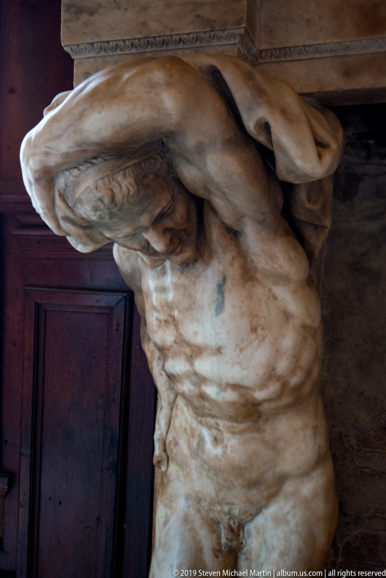 Weathered pale stone atlante with raised arms and bent posture supporting an overhead architectural element beside a dark door.