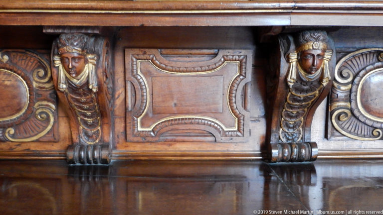 Polished carved wood panel with two female caryatid-like supports flanking a recessed rectangular decorative field.