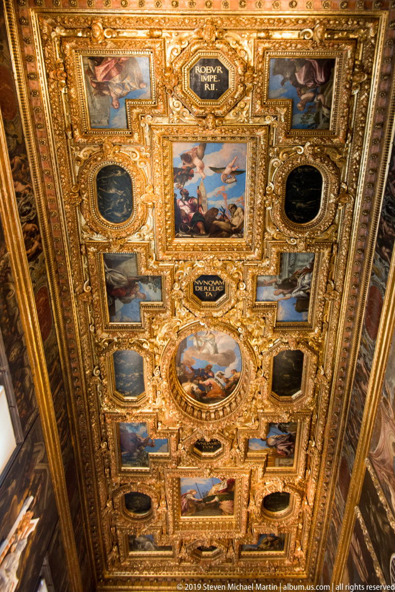 Oblique view along a richly gilded coffered ceiling filled with framed paintings, oval panels, and dark inscription cartouches.
