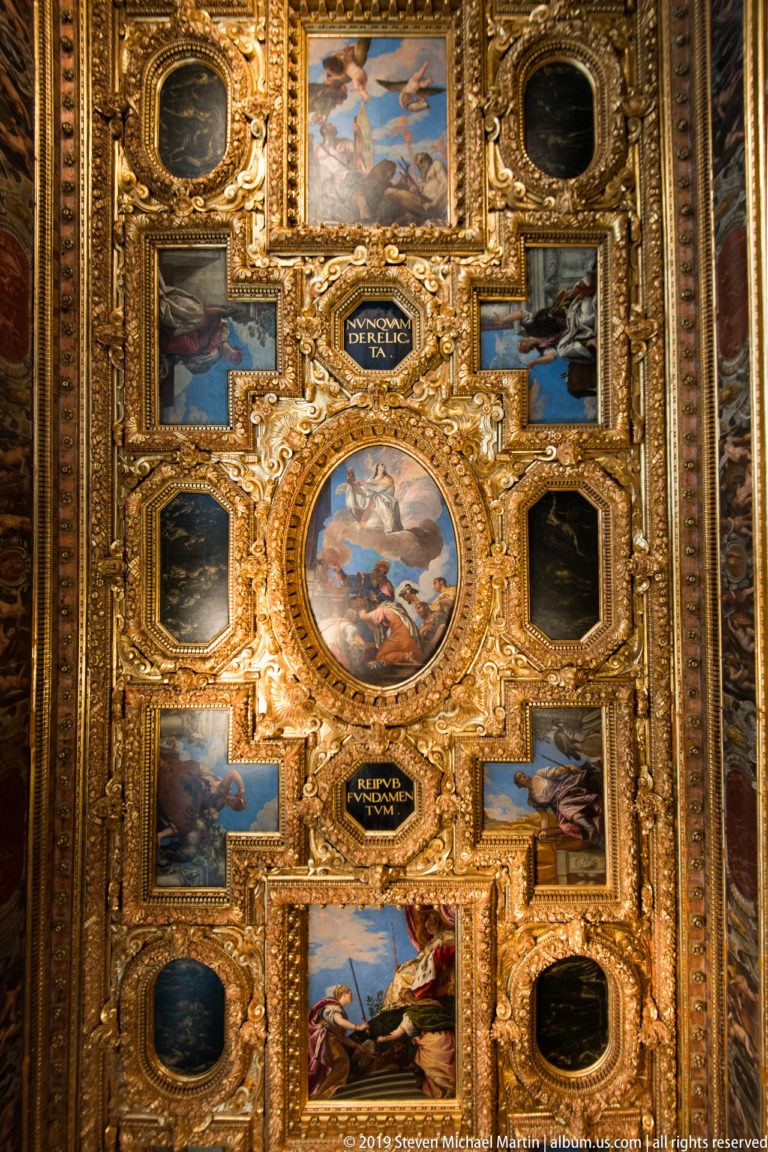 Gilded coffered ceiling with a central oval painting, smaller painted panels, dark cartouches, and dense carved ornament.