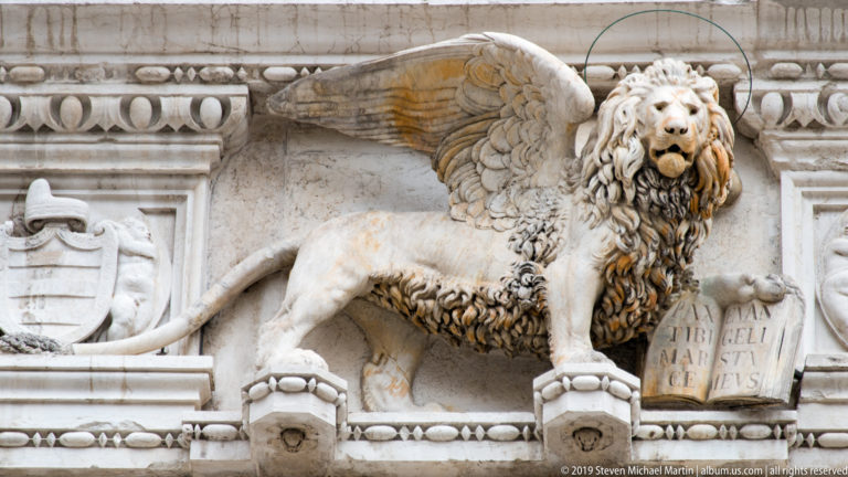 Carved winged lion with a large mane stands beside an open inscribed book on an ornate pale-stone façade.