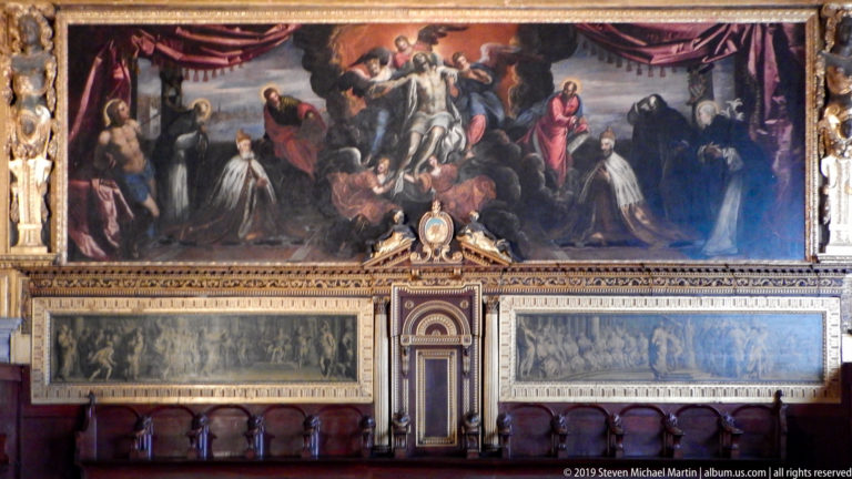 Large historical painting above carved wooden seating, pale relief panels, and an ornate architectural centerpiece in a ceremonial room.